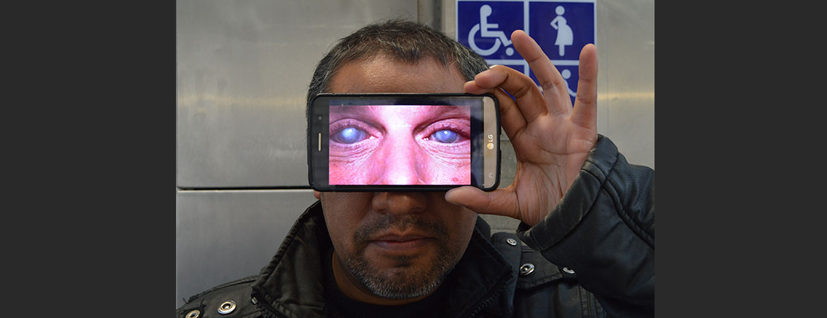 Hombre sosteniendo celular en frente de los ojos a modo de lentes, en el celular hay una imagen de unos ojos con nubosidad haciendo parecer que son los ojos del hombre, al fondo se observa el cartel con los símbolos de discapacidad